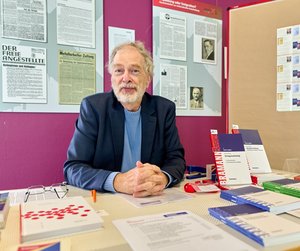 Klaus Bramann, sitzt an einem Schreibtisch, der mit verschiedenen Broschüren, Büchern und Informationsmaterialien bedeckt ist. Hinter der Person befindet sich eine lila Wand mit gerahmten Zeitungsartikeln und einem schwarzen Brett, das mit Mitteilungen und Dokumenten gefüllt ist.